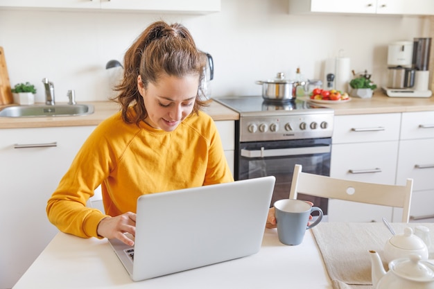Jovem mulher sentada na cozinha em casa, trabalhando, usando um laptop