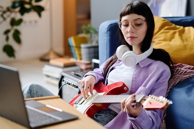 Jovem mulher sentada na frente do laptop e tocando violão ela gravando sua música no computador