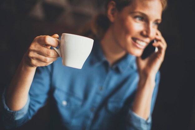 Jovem mulher sentada no café, usando telefone e bebendo café