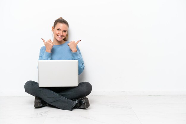 Foto jovem mulher sentada no chão com um laptop, fazendo gestos de polegar para cima e sorrindo