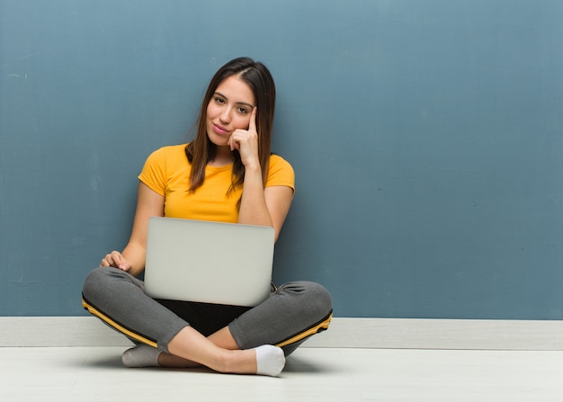 Jovem mulher sentada no chão com um laptop pensando em uma ideia