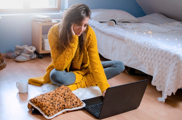 Jovem mulher sentada no chão no quarto dela, usando o laptop, olhando para a tela feliz em se comunicar com os amigos