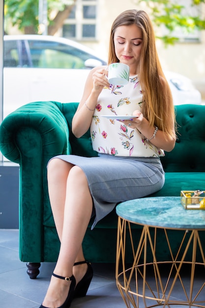 Jovem mulher sentada no sofá, bebendo chá da xícara em um escritório