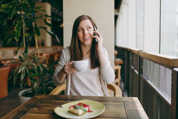 Jovem mulher sentada sozinha na cafeteria à mesa com uma xícara de cappuccino, bolo, relaxante no restaurante durante o tempo livre. Jovem mulher falando no celular, descansando no café. Conceito de estilo de vida.