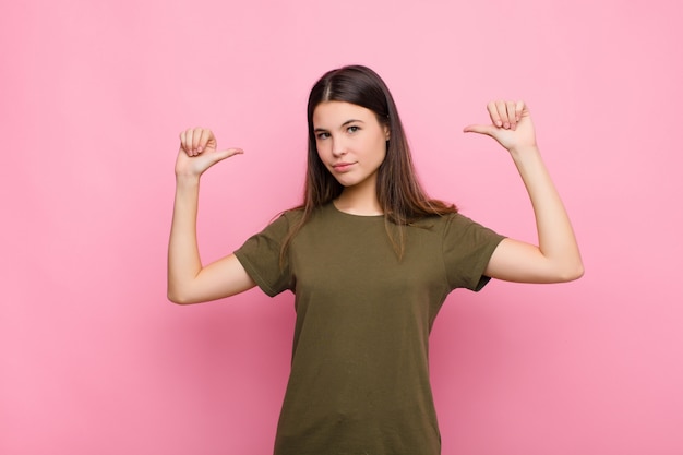 Jovem, mulher, sentindo-se orgulhoso, arrogante e confiante, olhando satisfeito e bem sucedido, apontando para si mesmo na parede rosa