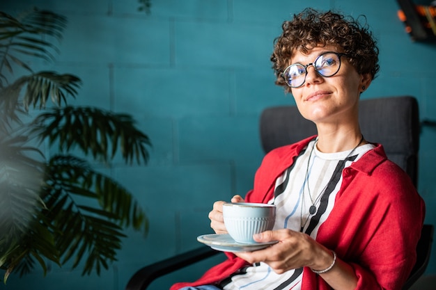 Jovem mulher serena e elegante com uma xícara de chá ou café relaxando no café na hora do almoço ou após um dia de trabalho