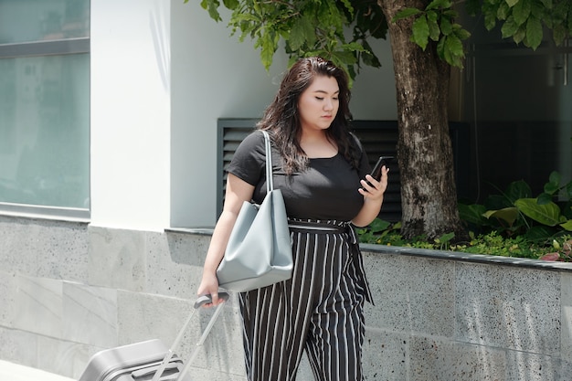 Jovem mulher séria com bagagem andando na rua e verificando mensagens de texto no smartohone
