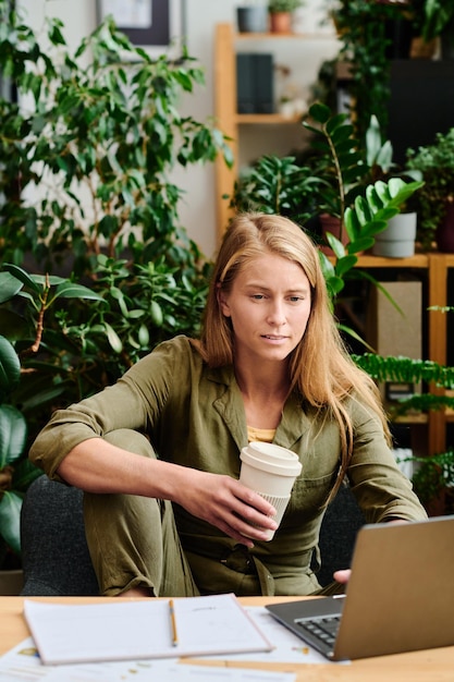 Jovem mulher séria com café sentado no local de trabalho em frente ao laptop