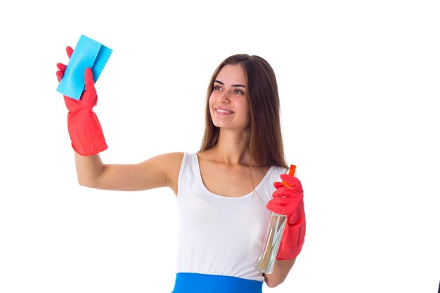 Jovem mulher séria na camisa branca e avental azul com luvas vermelhas usando espanador e detergente