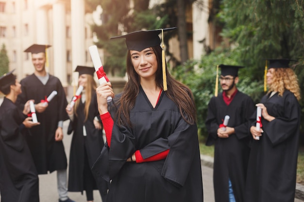 Jovem mulher séria no dia da formatura na universidade em pé com grupo multiétnico de pessoas. Educação, qualificação e conceito de vestido.