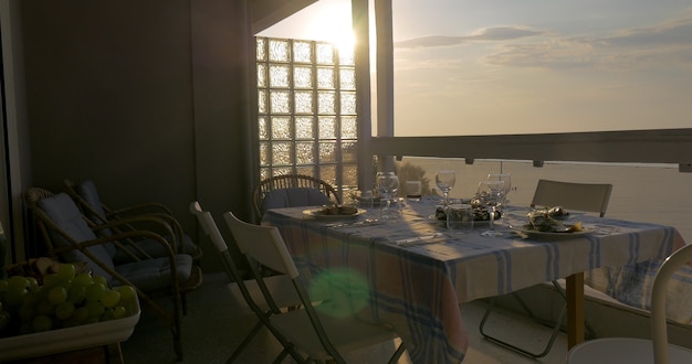 Jovem mulher servindo o jantar na varanda com vista para o mar ao pôr do sol. Pratos de peixe, copos e talheres na mesa