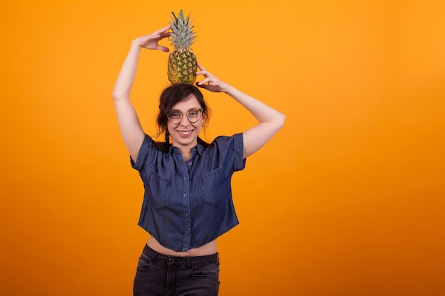 Jovem mulher sexy feliz sorrindo com óculos segurando um saboroso abacaxi na cabeça no estúdio sobre fundo amarelo. alegre mulher segurando deliciosos ananás.