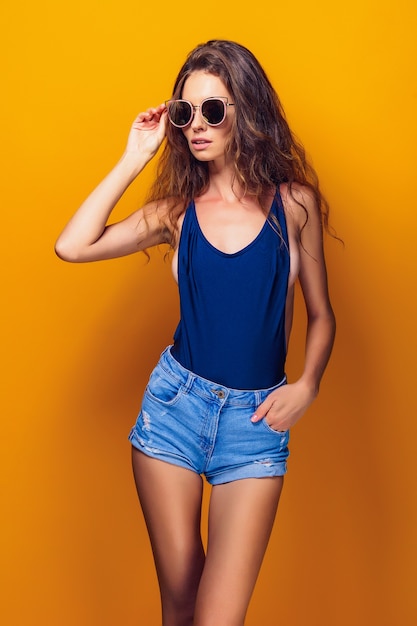 Jovem mulher sexy vestida de maiô e shorts jeans azul, óculos escuros posando no amarelo.