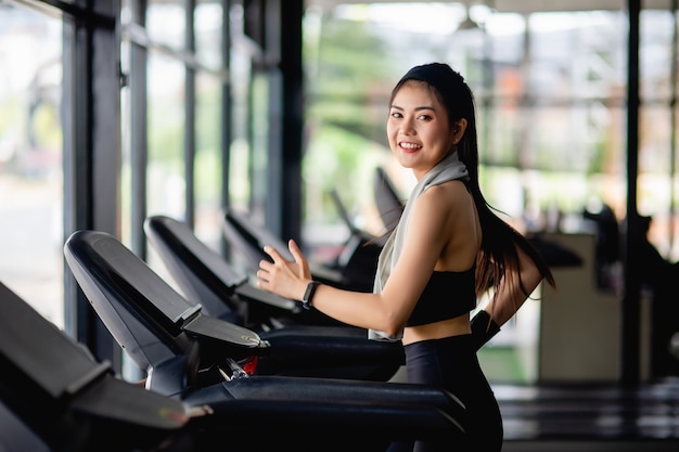 Jovem mulher sexy vestindo roupas esportivas, tecido à prova de suor e smartwatch correndo na esteira para se exercitar em uma academia moderna, sorria, copie o espaço