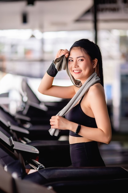 Jovem mulher sexy vestindo roupas esportivas, tecido à prova de suor e smartwatch usando uma toalha, limpe o suor na testa durante o treino em uma academia moderna, sorria, copie o espaço