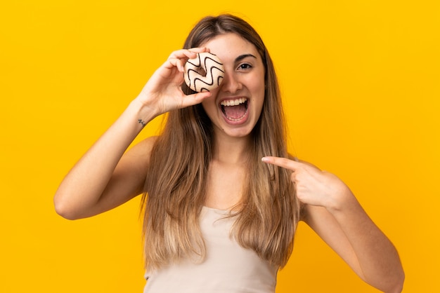 Jovem mulher sobre fundo amarelo isolado, segurando uma rosquinha e feliz