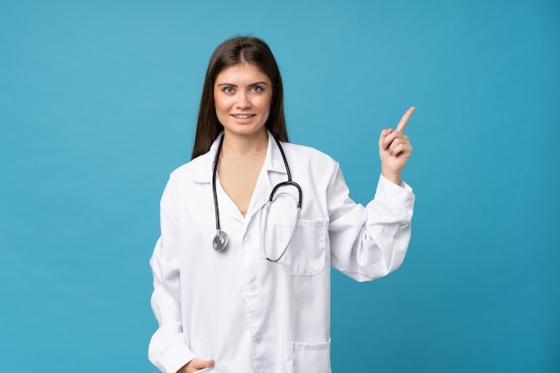 Jovem mulher sobre fundo azul isolado com vestido de médico e apontando o lado