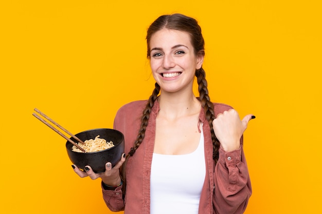 Jovem mulher sobre parede amarela isolada, apontando para o lado para apresentar um produto enquanto segura uma tigela de macarrão com pauzinhos