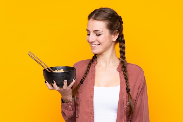 Foto jovem mulher sobre parede amarela isolada com expressão feliz, mantendo uma tigela de macarrão com pauzinhos