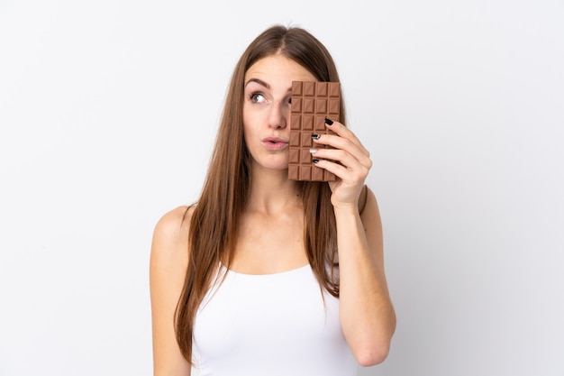 Foto jovem mulher sobre parede branca isolada, tomando um comprimido de chocolate e surpreendeu