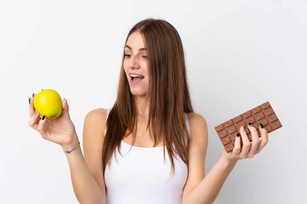 Jovem mulher sobre parede branca isolada, tomando uma tablete de chocolate em uma mão e uma maçã na outra