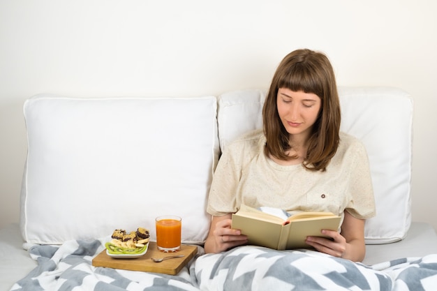 Jovem mulher sorri e lê livro na cama e toma café da manhã