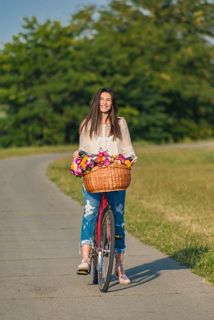 Jovem mulher sorridente anda de bicicleta com uma cesta cheia de flores na zona rural