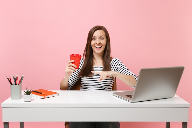 Jovem mulher sorridente apontando o dedo indicador para uma xícara de café ou chá, trabalhando em um projeto, sente-se no escritório com o laptop do pc