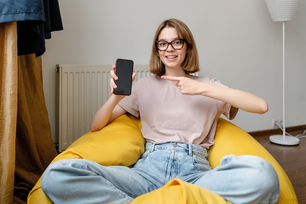 Jovem mulher sorridente apontando para um celular no apartamento usando uma camiseta azul jeans rosa e óculos