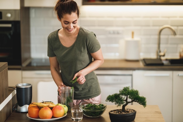 Jovem mulher sorridente colocando uma variedade de legumes em um liquidificador e fazendo um smoothie saudável em casa.