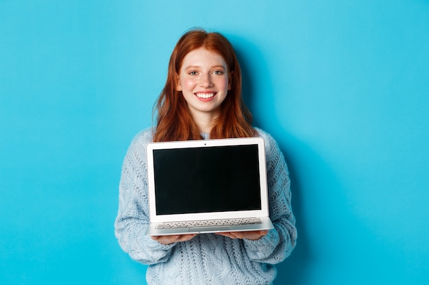 Jovem mulher sorridente com cabelo ruivo e sardas mostrando a tela do computador, segurando o laptop e demonstrar a promoção online, em pé sobre um fundo azul.