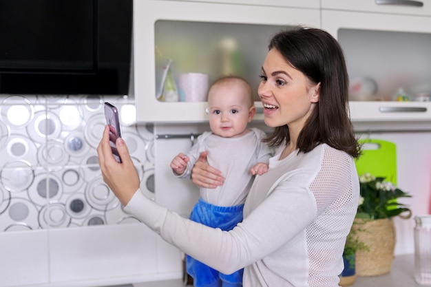 Jovem mulher sorridente com criança nos braços fazendo videochamada usando o smartphone em casa na cozinha. Bate-papo online com amigos, parentes. Tecnologia, conceito de estilo de vida à distância