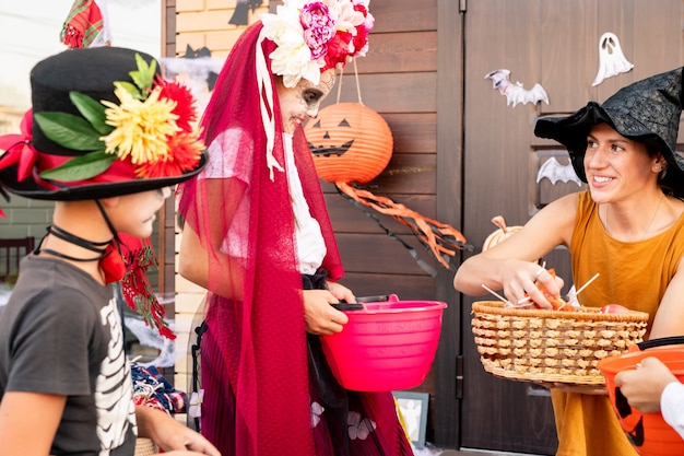 Jovem mulher sorridente com vestido amarelo e chapéu preto de bruxa, olhando para uma linda garota alegre de halloween com uma cesta e dando a ela alguns doces