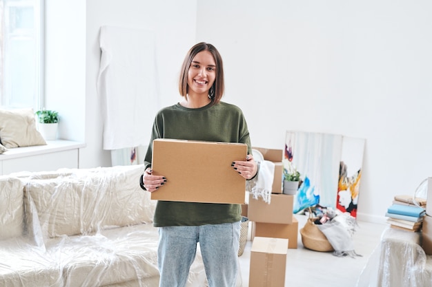 Jovem mulher sorridente em trajes casuais segurando uma caixa de papelão enquanto está de pé na sala de estar de um apartamento ou casa nova