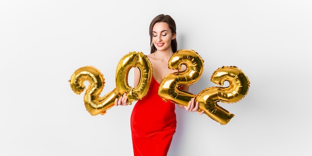 Jovem mulher sorridente em um vestido de cocktail vermelho com maquiagem brilhante, comemorando o ano novo e segurando golde.