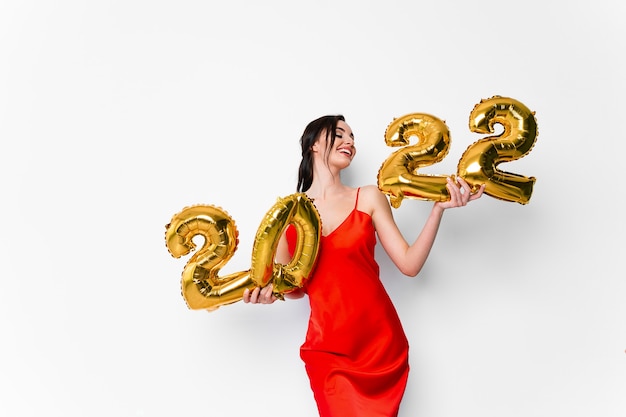Jovem mulher sorridente em um vestido de cocktail vermelho com maquiagem brilhante, comemorando o ano novo e segurando golde.