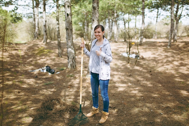 Jovem mulher sorridente, limpando com um ancinho para a coleta de lixo e aparecendo o polegar no parque cheio de lixo. Problema de poluição ambiental