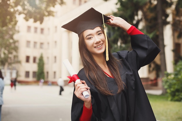 Jovem mulher sorridente no dia da formatura na universidade com diploma. Educação, qualificação e conceito de vestido.