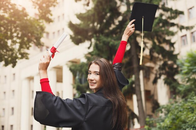 Jovem mulher sorridente no dia da formatura na universidade com diploma. Educação, qualificação e conceito de vestido.