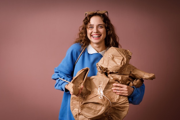 Jovem mulher sorridente positiva recebe um presente surpresa e segurando nas mãos um monte de presentes embrulhados em papel artesanal bege rústico chapéu sapatos secador de cabelo brinquedo macio e óculos