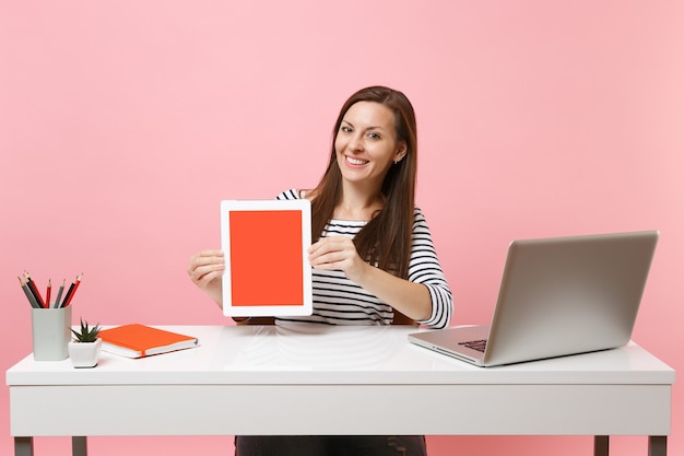 Jovem mulher sorridente segurando um computador tablet com a tela em branco e vazia, sente-se no trabalho na mesa branca com um laptop pc contemporâneo