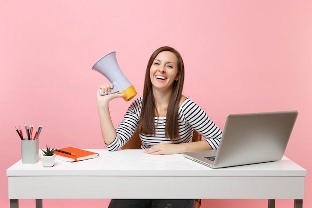 Jovem mulher sorridente segurando um megafone enquanto está sentado e trabalhando em um projeto na mesa branca no escritório com o laptop do pc