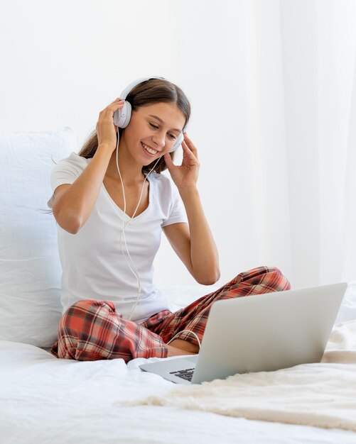 Jovem mulher sorridente, sentada na cama do quarto, ouvindo música ou assistindo filme