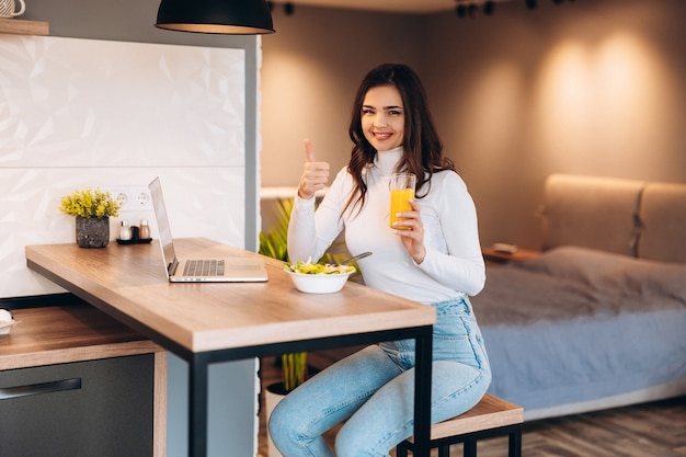 Jovem mulher sorridente tomando café da manhã na cozinha, ela está se conectando a um laptop e bebendo um suco de laranja saudável.