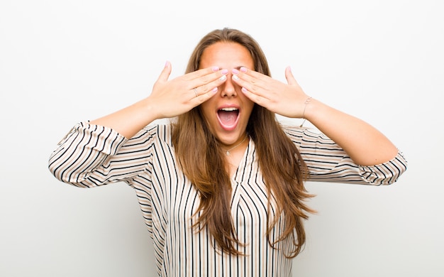 Foto jovem mulher sorrindo e se sentindo feliz, cobrindo os olhos com as duas mãos e esperando a surpresa inacreditável em branco