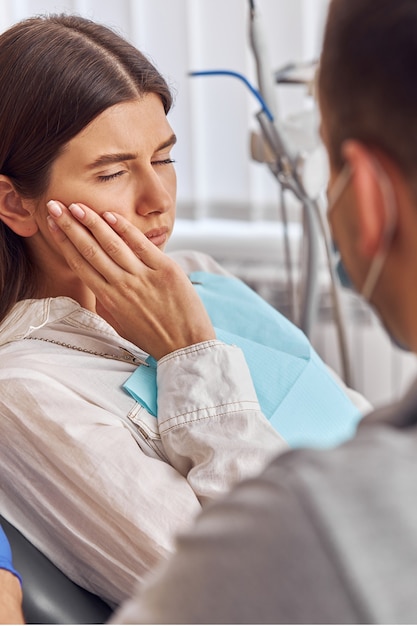 Foto jovem mulher tendo dor de dente, sentada em uma cadeira odontológica na clínica. paciente do sexo feminino com terrível dor de dente visitando o dentista.