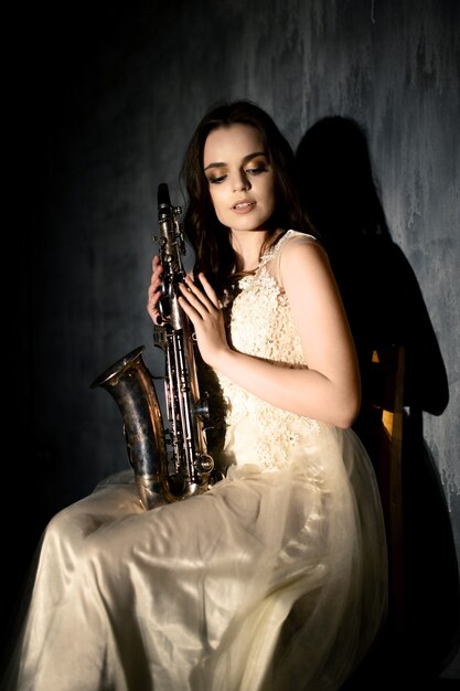 Jovem mulher toca saxofone. Menina com um vestido branco fofo posando com um instrumento musical