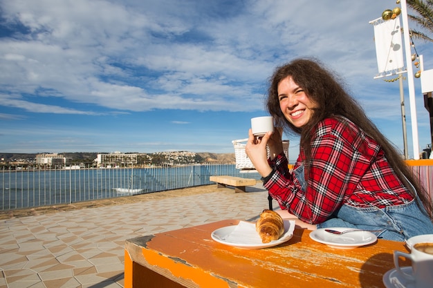 Jovem mulher tomando café da manhã com croissant e café no café na rua em frente ao mar.