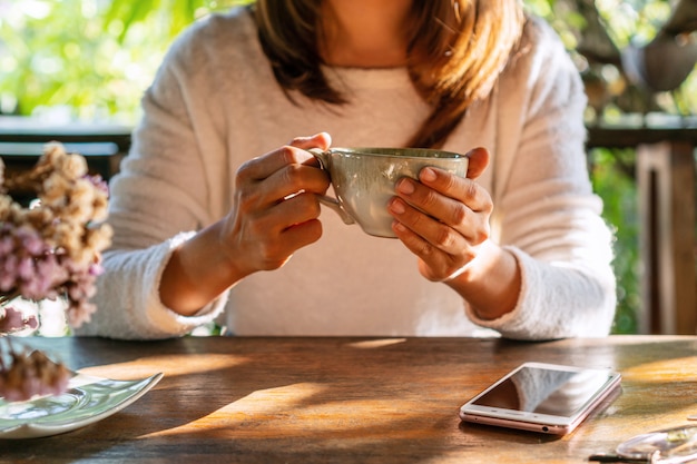 Jovem mulher tomando café na mesa de madeira com telefone inteligente no café durante o tempo livre.