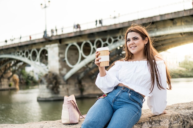 Jovem mulher tomando um café sentada à beira do rio em Sevilha, Espanha Menina sorridente, aproveitando o verão